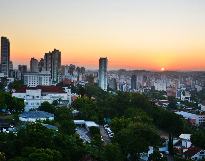 Foto que ilustra matéria sobre bairros de Novo Hamburgo mostra uma vista aérea da cidade com o sol se pondo no horizonte ao fundo. A imagem mostra, em primeiro plano, algumas árvores e casas e mais ao centro e à esquerda alguns prédios.