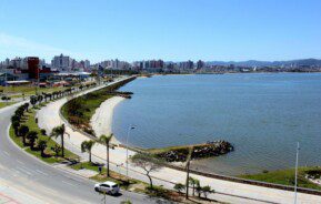 Foto que ilustra matéria sobre bairros de São José (SC) mostra um pedaço da orla da cidade, com o espelho d’água da baía à direita, a via pública à esquerda e prédios ao fundo.