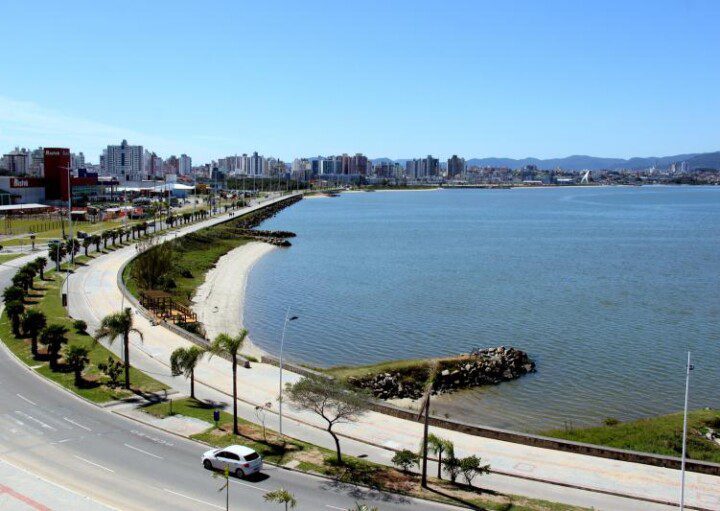 Foto que ilustra matéria sobre bairros de São José (SC) mostra um pedaço da orla da cidade, com o espelho d’água da baía à direita, a via pública à esquerda e prédios ao fundo.