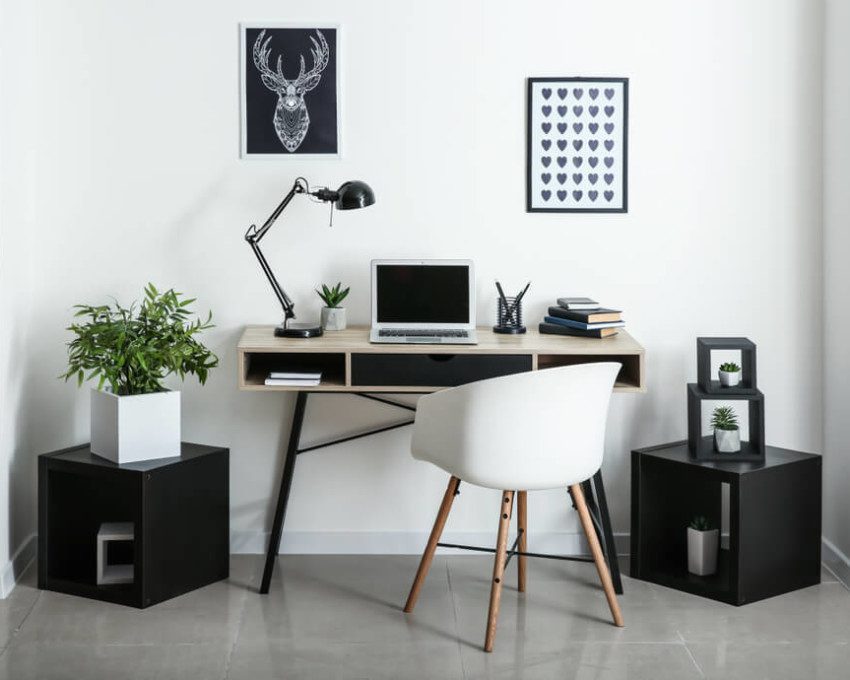 Foto que ilustra matéria sobre decoração de escritório moderno mostra uma mesa de computador encostada em uma parede branca com dois pequenos quadros acima. Em cima da mesa há um notebook, uma luminária e materiais de escritório, como canetas e papéis. Dos dois lados da mesa há dois nichos pretos em forma de cubo no chão. O da esquerda tem um vaso com uma planta.