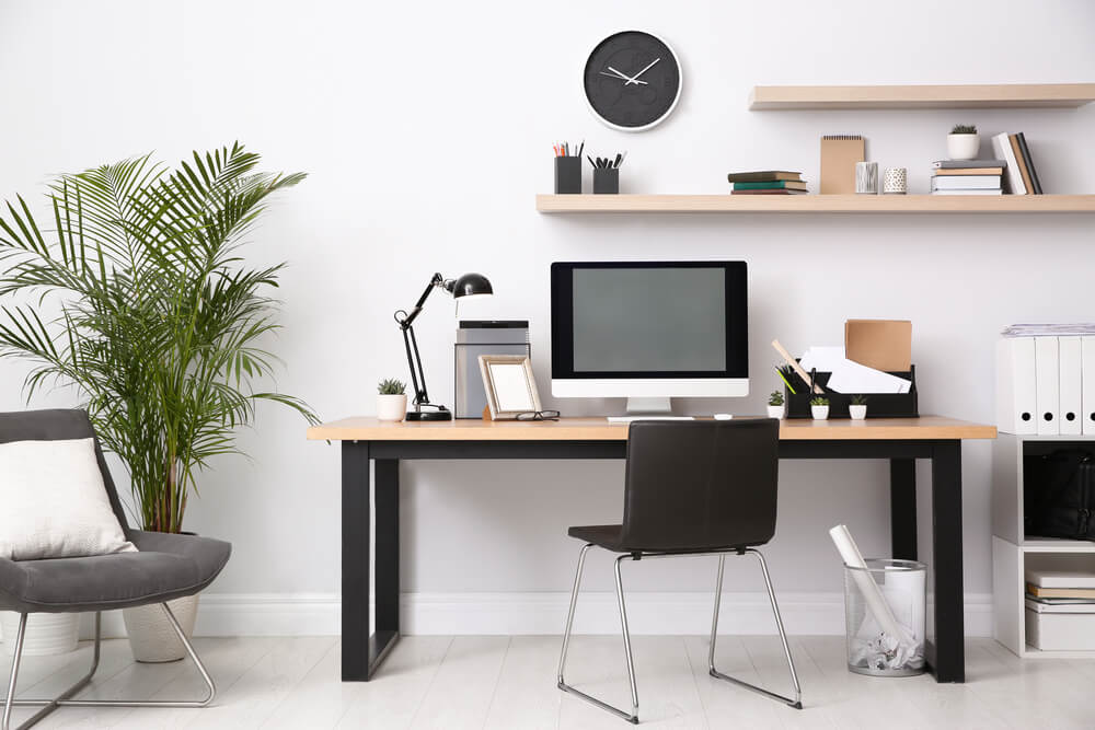 Foto que ilustra matéria sobre decoração de escritório moderno mostra um posto de trabalho em casa, com uma mesa com tampo de madeira clara e pés pretos, uma cadeira preta com pés cromados. Acima da mesa, há um monitor de computador e outros materiais de trabalho.