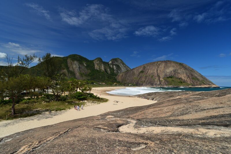 Foto que ilustra matéria sobre o que fazer em Niterói mostra uma vista panorâmica de um trecho da praia de Itacoatiara com uma pequena facha de areia, um pedaço das ondas do mar aparecendo e um grande maciço de pedra ao fundo em um dia de céu azul