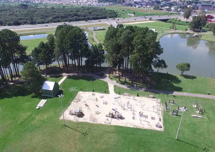 Foto que ilustra matéria sobre parques em São José dos Pinhais mostra a visão aérea do parque municipal da cidade, com um espaço de areia dentro de um gramado, onde se encontram aparelhos de ginástica, árvores e um lago mais ao fundo.