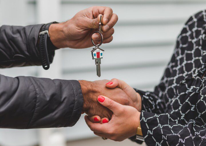 Imagem mostra mão masculina e feminina apertando, e uma das mãos segura uma chave de casa. Imagem disponível no Pexels