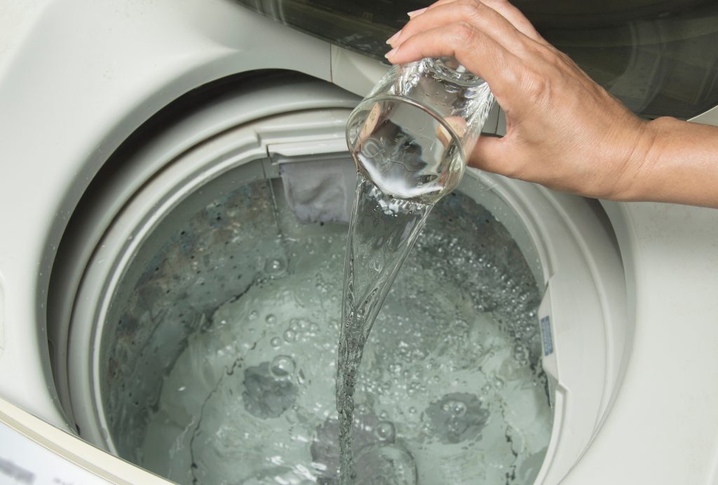 Imagem de uma pessoa adicionando um líquido transparente dentro de uma máquina para ilustrar matéria sobre como limpar máquina de lavar com vinagre