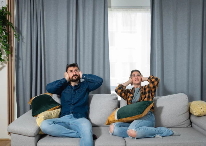 Imagem de um jovem casal, composto por um homem e uma mulher, sentado no sofá de casa olhando para cima com expressão de incômodo e com as mãos nos ouvidos para ilustrar matéria sobre perturbação do sossego