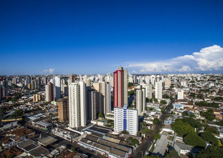 Foto que ilustra matéria sobre bairros de Uberlândia mostra uma visão do alto da cidade em um dia ensolarado de céu azul e poucas nuvens, com prédios muito altos no centro da imagem, algumas casas no canto inferior esquerdo e uma região arborizada no canto inferior direito.