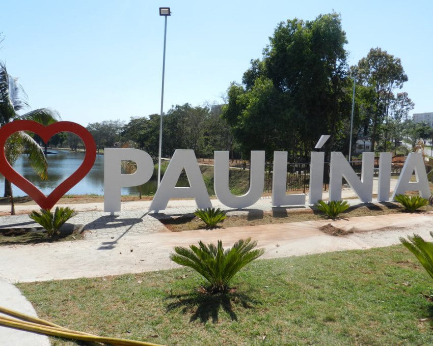 Foto que ilustra matéria sobre como chegar em Paulínia mostra uma imagem de um letreiro em um parque com um coração vermelho e o nome da cidade escrito em branco. Atrás, aparecem um lago, árvores e um céu azul claro.