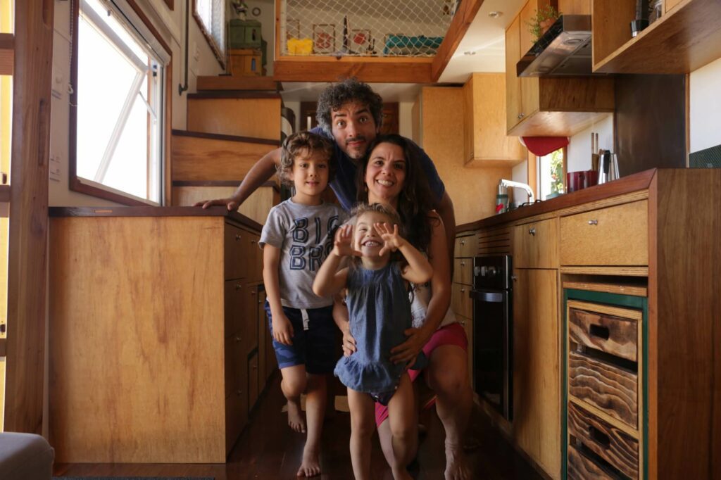 No pequeno corredor de uma Tiny House, um homem e uma mulher - Robson e Isabel - com seu casal de filhos posam sorridentes.