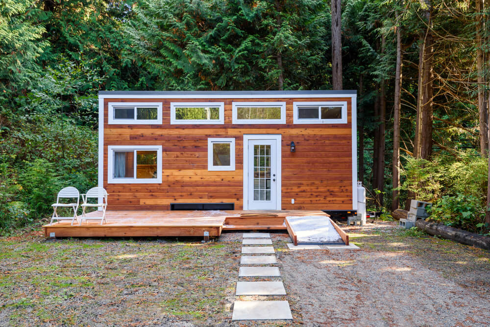 Foto que ilustra matéria sobre tiny house mostra uma pequena casa em formato retangular, com muitas árvores atrás. 