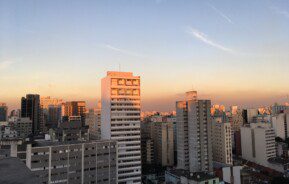 A imagem mostra a vista com prédios e casas no fim de tarde com o pôr do sol.