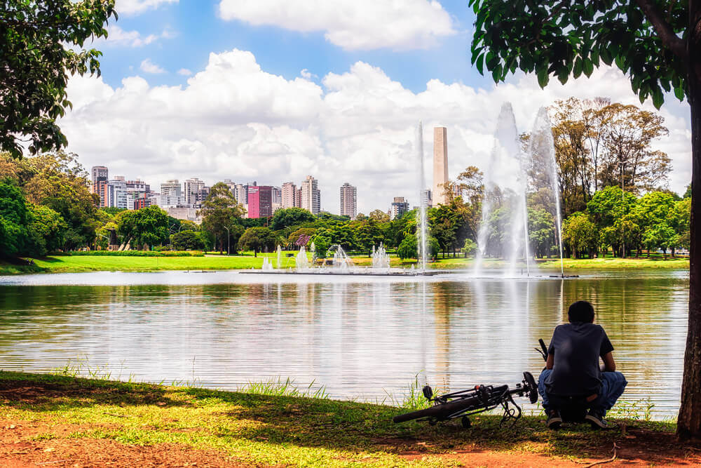 Foto que ilustra matéria sobre o que fazer em SP mostra um homem de costas, agachado, com sua bicicleta deitada no chão ao lado enquanto ele contempla um grande lago