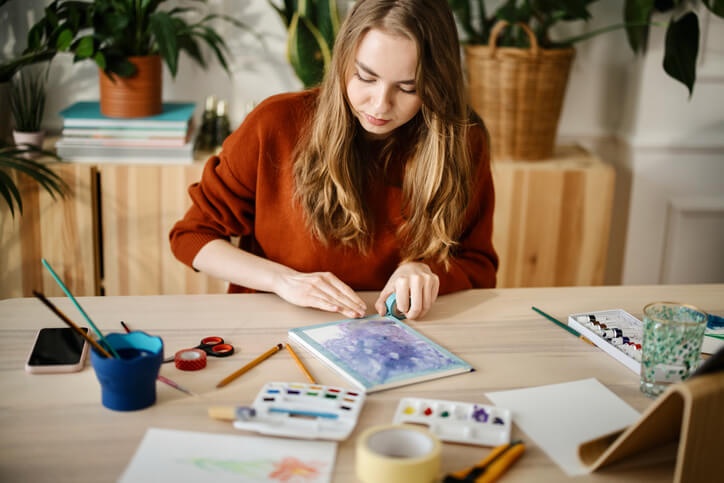 Foto que ilustra matéria sobre o que fazer em casa mostra uma mulher fazendo um pequeno quadro em aquarela na mesa de sala com tintas em cima da mesa