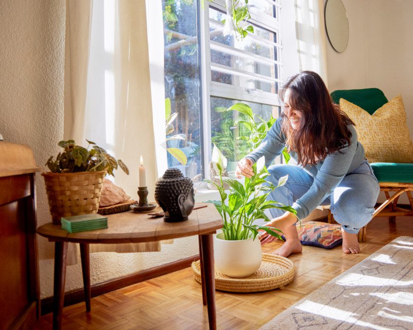 Imagem de uma mulher cuidando de uma planta ao lado de uma mesa com vela e um buda decorativo.