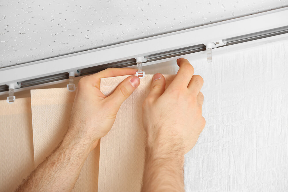 Foto que ilustra matéria sobre tipos de cortina mostra as mãos de um homem em detalhe instalando uma cortina em um trilho preso ao teto.