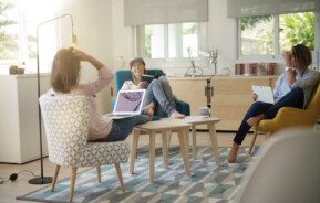 Foto que ilustra matéria sobre coliving mostra um grupo de três jovens sentadas em uma sala de estar. Elas conversam e riem. Duas delas têm notebooks em seus colos, enquanto a terceira tem um livro aberto nas mãos