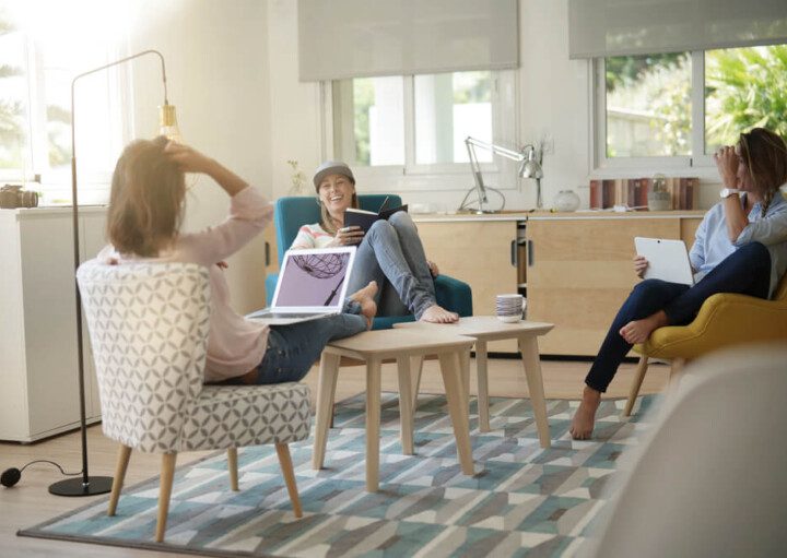 Foto que ilustra matéria sobre coliving mostra um grupo de três jovens sentadas em uma sala de estar. Elas conversam e riem. Duas delas têm notebooks em seus colos, enquanto a terceira tem um livro aberto nas mãos