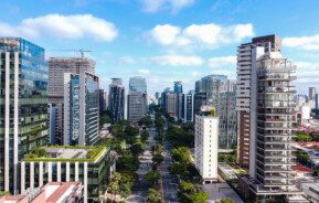 Foto de uma cidade mostrada de cima. Nela há uma grande avenida e diferentes prédios ao redor.