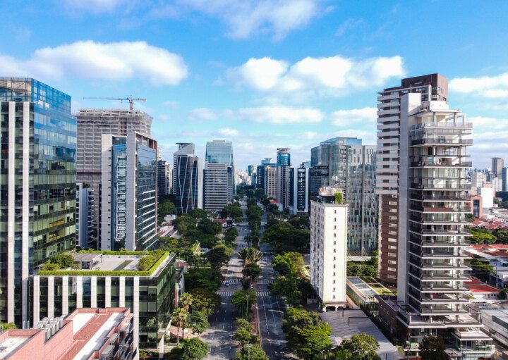 Foto de uma cidade mostrada de cima. Nela há uma grande avenida e diferentes prédios ao redor.