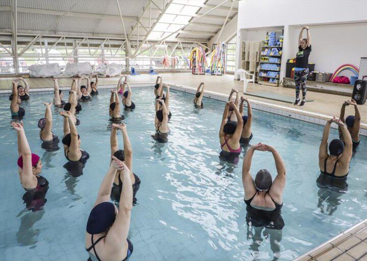 Foto que ilustra matéria sobre o programa Curitiba em Movimento mostra uma piscina com várias mulheres dentro com os braços levantados acima da cabeça fazendo alongamento, enquanto são orientadas por uma instrutora de pé na borda, do lado de fora.