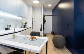 Foto mostra o interior de um moderno microapartamento, com um espaço com bancada e armários de cozinha brancos, integrados a uma pequena mesa. Do lado oposto, uma parede azul com uma porta. E no meio, ao fundo, a porta de entrada do imóvel.