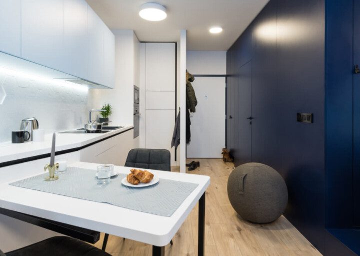 Foto mostra o interior de um moderno microapartamento, com um espaço com bancada e armários de cozinha brancos, integrados a uma pequena mesa. Do lado oposto, uma parede azul com uma porta. E no meio, ao fundo, a porta de entrada do imóvel.