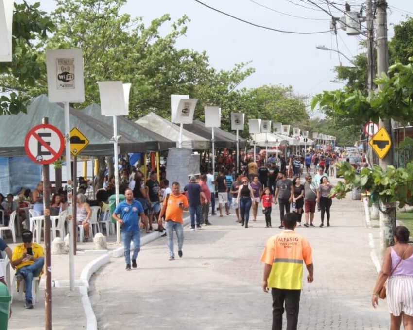 Foto que ilustra matéria sobre a Praia das Pedrinhas mostra a rua adjascente repleta espaços cobertos por toldos onde muitas pessoas circulam ou estão sentadas