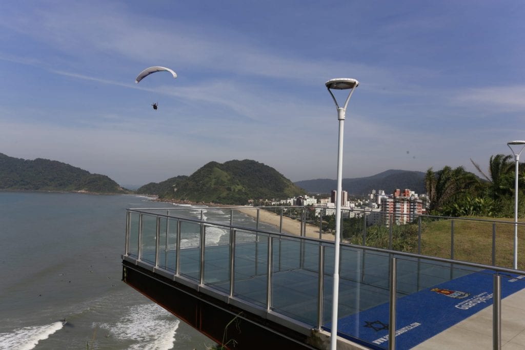 Imagem de um mirante com estrutura toda em vidro e no fundo o mar com pessoas praticando voo livre.