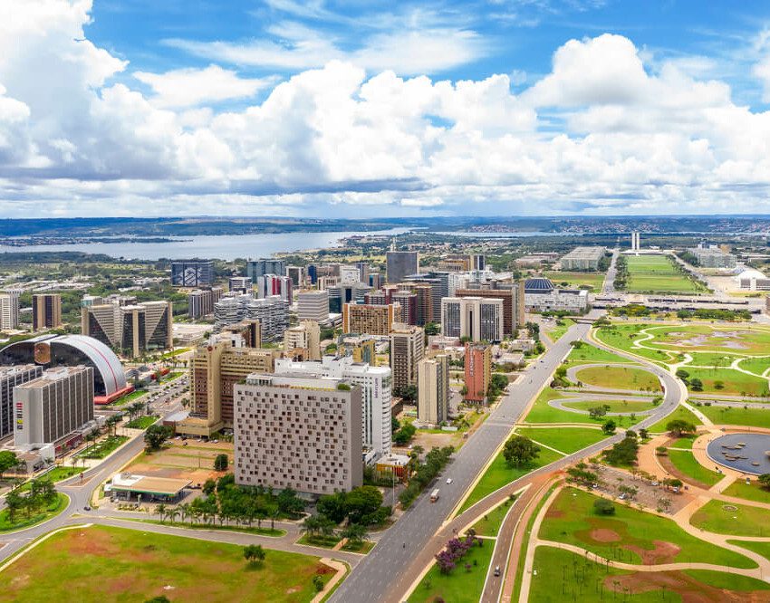 Foto que ilustra matéria sobre o Eixo Monumental mostra a região do alto, com alguns prédios à esquerda, uma avenida no meio e um céu azul com nuvens ao fundo