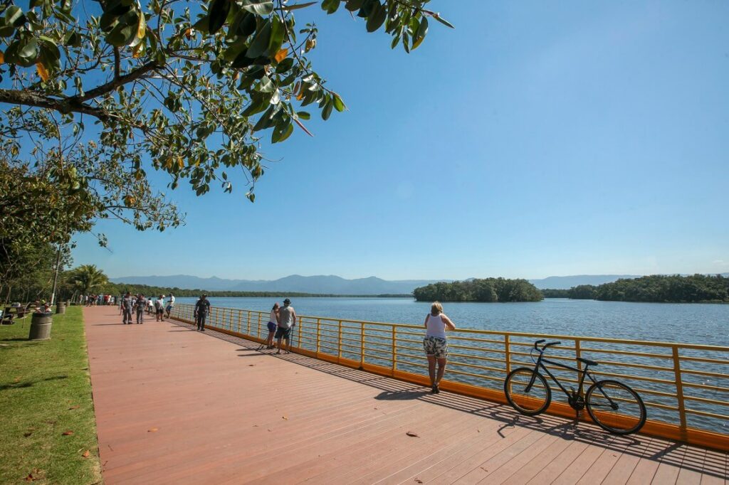 Foto que ilustra matéria sobre parques em Praia Grande um grande deck com grade amarela com vista para um grande espelho d'água, com pessoas caminhando e contemplando a paisagem.