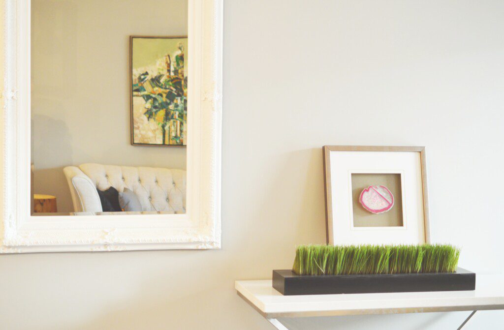 Aparador com quadro e vaso de planta e ao lado um espelho com moldura branca e clássica. 