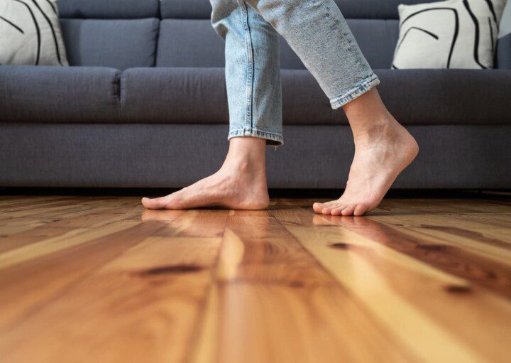 Uma mulher de pés descalços caminha sobre um piso de taco, é possível ver um sofá no fundo da foto.