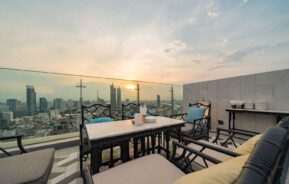 Foto que ilustra matéria sobre rooftop mostra uma área externa no topo de um prédio com uma mesa central, algumas poltronas em volta e uma vista da cidade ao cair da tarde