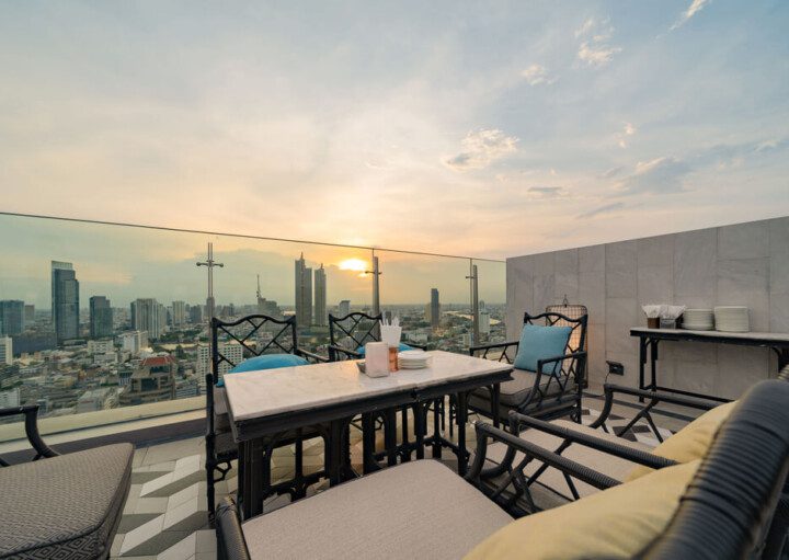 Foto que ilustra matéria sobre rooftop mostra uma área externa no topo de um prédio com uma mesa central, algumas poltronas em volta e uma vista da cidade ao cair da tarde