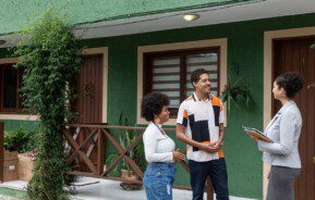 Foto que ilustra matéria sobre casa de vila mostra um casal sorridente conversando com uma corretora de imóveis em frente a uma casa verde