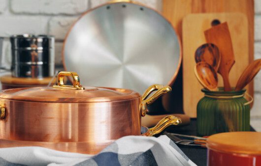 Imagem de uma panela de cobre com utensílios de madeira perto, como talheres e tábuas, em cima de uma mesa estampada com uma toalha quadriculada