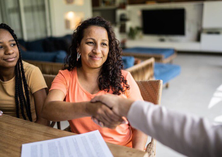 Duas mulheres negras, provavelmente mãe e filha, fecham um contrato de financiamento apertando a mão de um consultor.