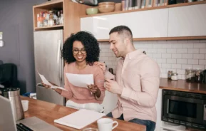 Imagem de um casal composto por um homem e uma mulher analisando as contas de casa sentados na mesa da cozinha para ilustrar matéria sobre alienação fiduciária