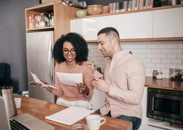 Imagem de um casal composto por um homem e uma mulher analisando as contas de casa sentados na mesa da cozinha para ilustrar matéria sobre alienação fiduciária