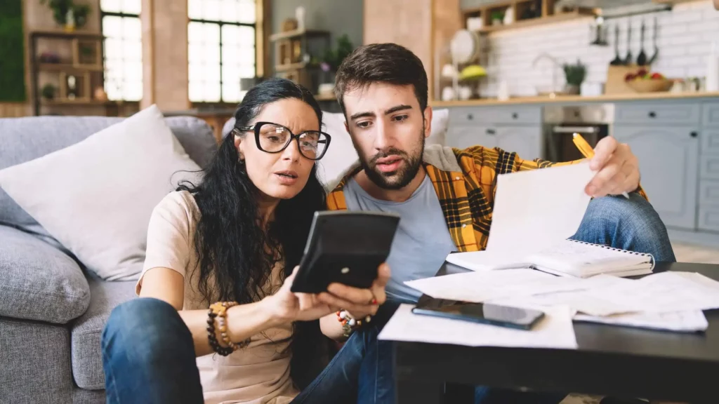 Imagem de um casal composto por um homem e uma mulher sentados no chão da sala de estar em frente ao sofá analisando documentos, papéis e contas da casa para ilustrar matéria sobre como tirar alienação fiduciária de imóvel