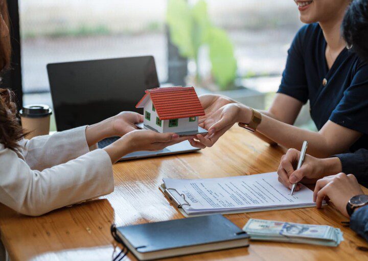 Duas pessoas assinam um contrato de compra conjunta de imóvel. Uma mulher, provavelmente de uma imobiliária, passa a miniatura de uma casa para um dos compradores.