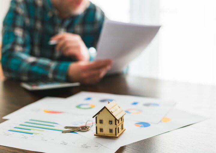 Miniatura de uma casa em primeiro plano, em cima de alguns documentos. Ao fundo há um homem lendo um documento.