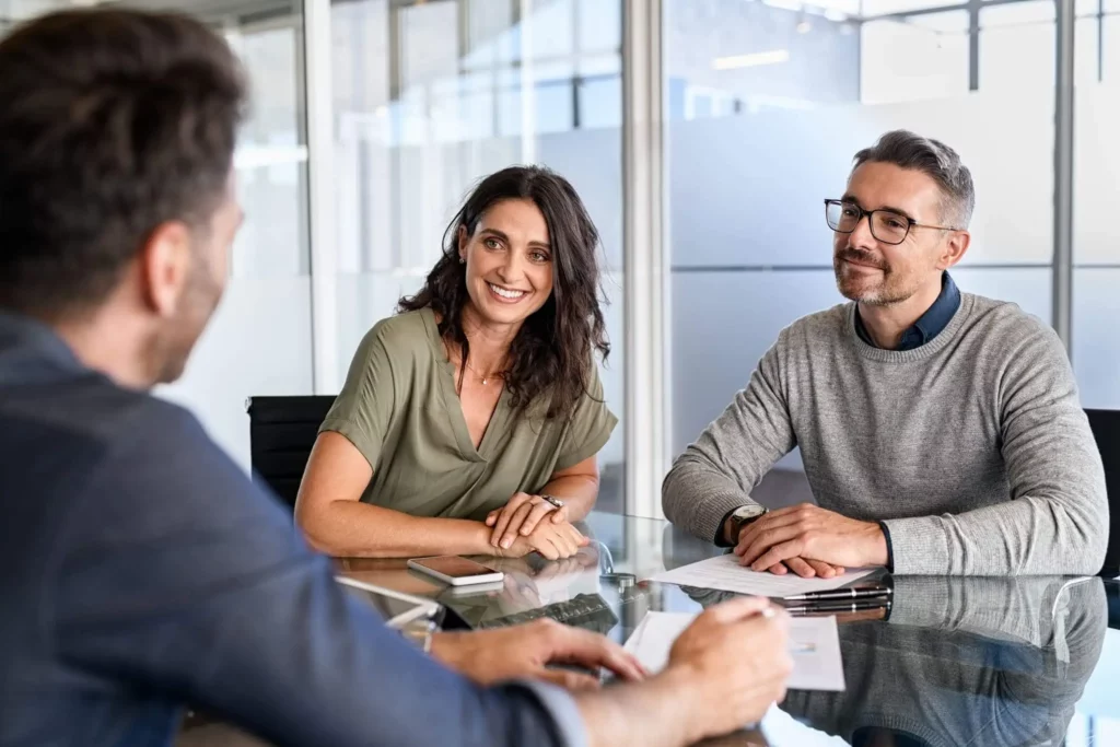 Imagem de um casal sentados composto por um homem e uma mulher sentados nas cadeiras de uma mesa de frente para um homem que aparece de costas para ilustrar matéria sobre o que significa alienação fiduciária