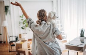 Foto que ilustra matéria sobre construir ou comprar imóvel novo mostra uma mulher vendo a sala de uma casa e segurando seu cachorro