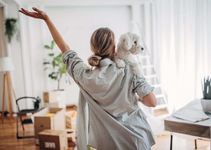 Foto que ilustra matéria sobre construir ou comprar imóvel novo mostra uma mulher vendo a sala de uma casa e segurando seu cachorro