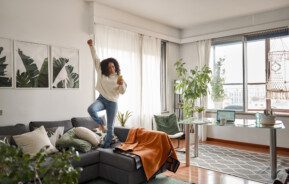 Imagem de uma jovem feliz e animada escutando música em cima do sofá, com um dos braços para cima, em um apartamento com estilo jovial, com uma janela à direita e plantas à esquerda, para decorar matéria sobre apartamento para solteiro