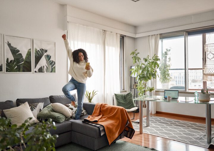 Imagem de uma jovem feliz e animada escutando música em cima do sofá, com um dos braços para cima, em um apartamento com estilo jovial, com uma janela à direita e plantas à esquerda, para decorar matéria sobre apartamento para solteiro