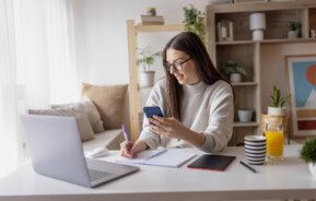 Imagem de uma mulher trabalhando em casa tomando notas no caderno e no celular para ilustrar matéria sobre como economizar dinheiro para comprar um apartamento