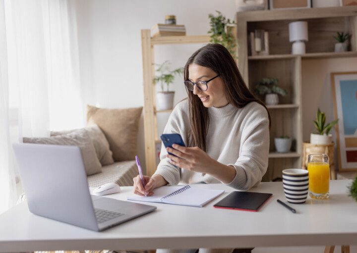 Imagem de uma mulher trabalhando em casa tomando notas no caderno e no celular para ilustrar matéria sobre como economizar dinheiro para comprar um apartamento