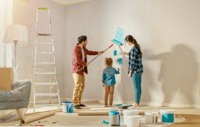 Foto de uma mulher, um homem e uma criança pequena pintando a parede de casa com um rolo pintura mergulhado em tinta azul para ilustrar matéria sobre como economizar na reforma da casa